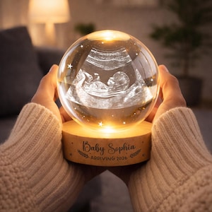 May include: A glowing crystal ball with a baby ultrasound image inside, resting on a wooden base. The base is engraved with the text "Baby Sophia ARRIVING 2026". The ball is lit from within, and surrounded by small star-shaped lights.
