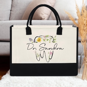 Peut inclure: Un sac fourre-tout en toile avec des poignées noires et un panneau inférieur noir. Le sac présente un motif floral autour d'une dent, avec le texte "Dr. Sandra M". Le sac est posé sur un tapis blanc.