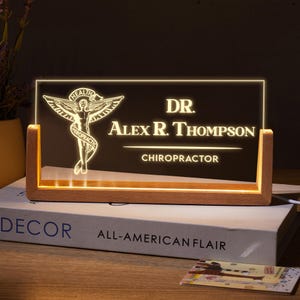 May include: A wooden sign with a lit up acrylic insert. The insert features a medical symbol of a staff with a snake wrapped around it. The text "DR. Alex R. Thompson, Chiropractor" is below the symbol.