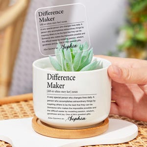 May include: A white ceramic pot with the text "Difference Maker" and a definition, holding a succulent plant. A clear acrylic sign with the same text and "Sophia" is inserted into the plant. The pot sits on a small wooden coaster.