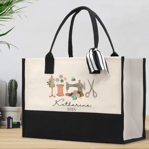 Peut inclure: Un sac fourre-tout en toile avec des poign&eacute;es noires et un panneau inf&eacute;rieur noir. La partie sup&eacute;rieure cr&egrave;me pr&eacute;sente une illustration &agrave; l'aquarelle d'outils de couture, dont une machine &agrave; coudre, des ciseaux et une bobine de fil. Le nom "Katherine 2025" est &eacute;crit en dessous.