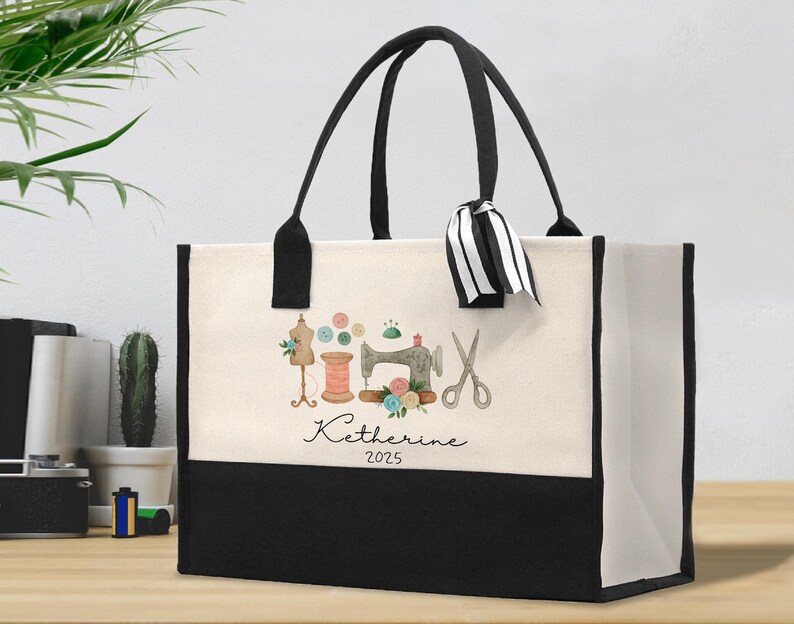 Peut inclure: Un sac fourre-tout en toile avec des poign&eacute;es noires et un panneau inf&eacute;rieur noir. La partie sup&eacute;rieure cr&egrave;me pr&eacute;sente une illustration &agrave; l'aquarelle d'outils de couture, dont une machine &agrave; coudre, des ciseaux et une bobine de fil. Le nom "Katherine 2025" est &eacute;crit en dessous.