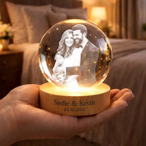 Op de afbeelding: Een heldere glazen bol met een foto van een stel erin, geplaatst op een houten voet. De voet is gegraveerd met de namen "Sadie & Kevin" en de datum "22.12.2012". De bol wordt van binnenuit verlicht, wat een warme gloed creëert.