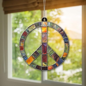 Puede incluir: Un adorno de vidrio emplomado en forma de signo de paz con un diseño abstracto colorido. El adorno está colgado en una ventana con vista a árboles verdes.