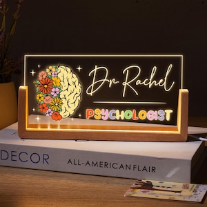 Könnte beinhalten: Ein Schild aus Holz mit einem leuchtenden Acryleinsatz. Der Einsatz zeigt ein buntes Gehirn mit Blumen und den Text "Dr. Rachel Psychologin".