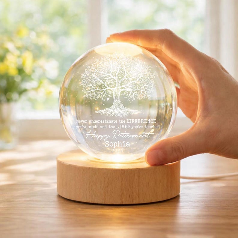 K&ouml;nnte beinhalten: Eine klare Glaskugel mit einem Baumdesign und den Worten "Never underestimate the DIFFERENCE you've made and the LIVES you've touched. Happy Retirement Sophia 2026" wird von einem Holzsockel beleuchtet.