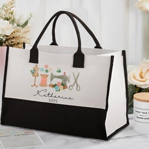 Peut inclure: Un sac fourre-tout blanc avec des bordures et des poign&eacute;es noires. Le sac pr&eacute;sente un motif aquarelle d'outils de couture, dont une machine &agrave; coudre, des ciseaux et du fil. Le nom "Katherine" et l'ann&eacute;e "2025" sont &eacute;galement imprim&eacute;s sur le sac.
