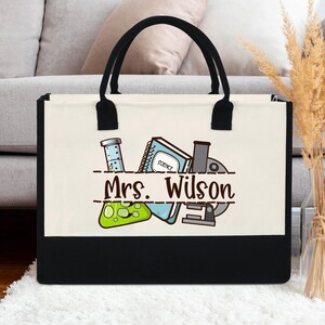 Peut inclure: Un sac fourre-tout en toile crème avec des poignées et un fond noirs. Le sac présente un motif sur le thème de la science avec un bécher, un microscope et un carnet, et le nom "Mrs. Wilson" est imprimé.