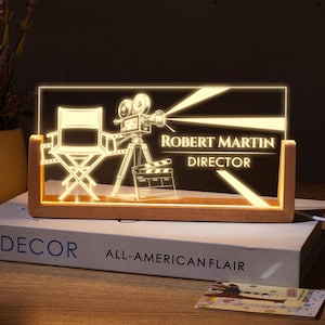 May include: A rectangular LED acrylic sign with a wooden base featuring an engraved director chair, a vintage film camera, and a film clapperboard. The text reads ROBERT MARTIN DIRECTOR in a glowing warm light against a dark background.