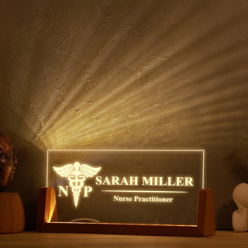 May include: A lighted desk nameplate with the name "SARAH MILLER" and the title "Nurse Practitioner" is displayed. The nameplate features a medical symbol and is set in a wooden base. The light shines from the nameplate.