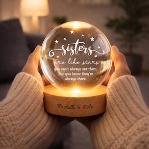 Könnte beinhalten: Ein kugelförmiges Nachtlicht aus Glas mit warmem Innenlicht, verziert mit dem weißen Text "sisters are like stars you can't always see them, but you know they're always there." und kleinen Sternsymbolen. Der Holzsockel ist mit "Michelle & Kate" graviert.