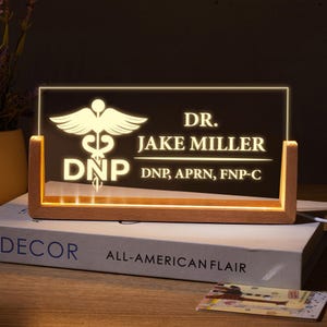 May include: A wooden sign with a lit up acrylic insert featuring a medical symbol, the text "DR. JAKE MILLER DNP, APRN, FNP-C", and a decorative wooden base.