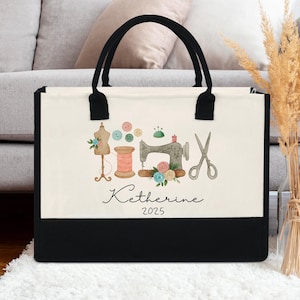 Peut inclure: Un sac fourre-tout en toile avec des poign&eacute;es noires et un panneau inf&eacute;rieur noir. Le panneau avant blanc cass&eacute; pr&eacute;sente une illustration &agrave; l'aquarelle d'outils de couture, dont une machine &agrave; coudre, des ciseaux et une bobine de fil. Le nom "Katherine" et l'ann&eacute;e "2025" sont &eacute;crits en dessous.