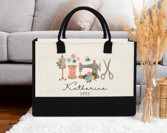 May include: A canvas tote bag with black handles and a black bottom panel. The off-white front panel features a watercolor illustration of sewing tools, including a sewing machine, scissors, and a spool of thread. The name "Katherine" and the year "2025" are written below the illustration.
