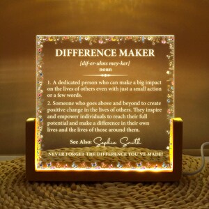 Könnte beinhalten: Eine beleuchtete Acryl-Plakette mit einer Definition des Wortes "Difference Maker" und dem Text "Vergiss nie den Unterschied, den du gemacht hast!" Die Plakette hat einen Holzrahmen und einen dekorativen Rand aus Schmetterlingen. Der Name "Sophia Smith" ist im Text enthalten.