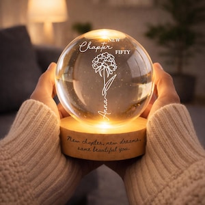 Puede incluir: Un globo de cristal transparente con un diseño floral y el nombre "Anna" grabado, apoyado sobre una base de madera. La base tiene la inscripción "New chapter, new dreams, same beautiful you." El globo está iluminado desde dentro.