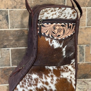 May include: A brown and white cowhide boot bag with a leather floral design on the front pocket. The bag has a zipper closure and a handle for carrying. The bag is designed to hold a pair of boots.