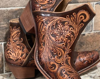 May include: A pair of brown leather ankle boots with intricate floral and scroll tooling. The boots have a square toe, stacked heel, and open sides. The leather is a rich brown with lighter tan accents.