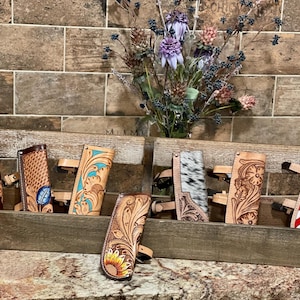 May include: A wooden display box holds several leather phone cases with intricate floral and geometric designs. One case features the American flag, while another showcases a sunflower. The cases are in various shades of brown and tan.