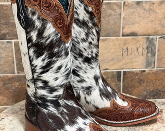 May include: A pair of tall western-style boots featuring a black and white cowhide pattern on the shaft and foot. The boots are accented with brown leather overlays on the toe and collar, both decorated with intricate floral scrollwork tooling.