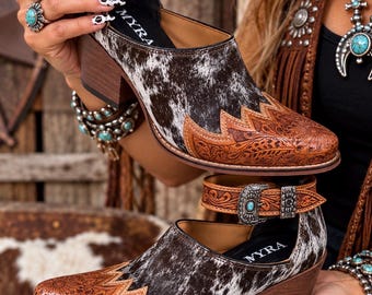 May include: A pair of ankle boots featuring a brown and white cowhide pattern, a tan leather toe cap with intricate floral tooling, a stacked wooden heel, and an adjustable ankle strap with a silver buckle and a small turquoise stone inlay.