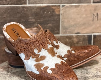 Peut inclure: Une paire de mules à enfiler en cuir marron avec des motifs floraux gravés et des empiècements en peau de vache blanche et marron. La semelle intérieure est beige avec le nom de marque MYRA en noir, dotée d'un talon en bois empilé.