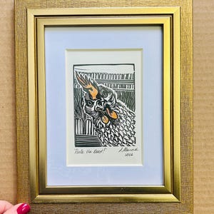 May include: A framed print with a black and white linocut of a rooster, with orange details. The artwork is set against a white mount and framed in a gold-coloured frame. The text "Rule the roost!" is visible.