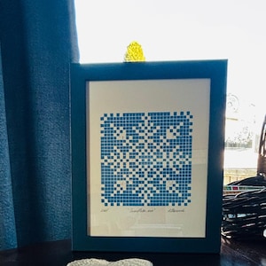 May include: A framed print with a blue and white pixelated snowflake design. The artwork is in a dark teal frame, on a dark surface. A white and pink crocheted coaster is in the foreground.