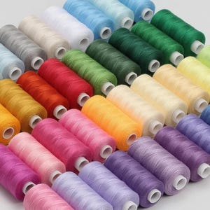 May include: A collection of sewing thread spools arranged in a grid, featuring a wide spectrum of colors including shades of red, orange, yellow, green, blue, purple, pink, white, gray, and cream, each with a white plastic core.