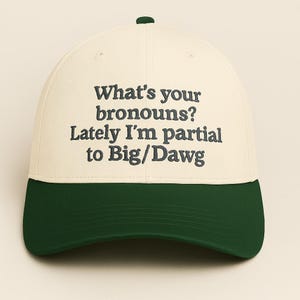Op de afbeelding: Een baseballpet met een crèmekleurige kroon en een donkergroene klep. De pet heeft de geborduurde tekst: "What's your pronouns? Lately I'm partial to Big/Dawg."