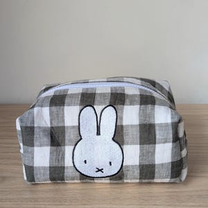 May include: A rectangular, zippered pouch with a black and white gingham pattern. The pouch features an embroidered white bunny face with black details. The zipper is white, and the pouch is sitting on a wooden surface.