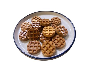 Puede incluir: Un plato blanco con borde azul contiene una pila de pequeños gofres redondos. Los gofres son de color marrón dorado con un patrón de cuadrícula cuadrada. El plato está sobre un fondo blanco.