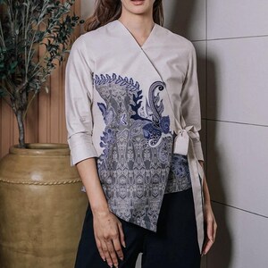 Op de afbeelding: Een crèmekleurige wikkel top met een blauw en grijs paisley design op de voorkant. De top heeft driekwart mouwen en een striksluiting in de taille. De top wordt gecombineerd met een zwarte broek.