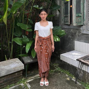 May include: A woman wearing a white blouse and a brown patterned skirt with white accents. She is standing in front of a green leafy plant and a white bench.