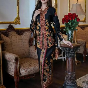 Puede incluir: Un vestido tradicional negro con un estampado floral en tonos rojos, naranjas y amarillos. El vestido presenta mangas largas y una falda a juego. El atuendo se usa con tacones plateados. El fondo incluye una silla decorativa y un jarr&oacute;n de rosas rojas.