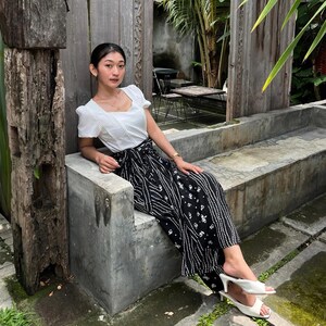 May include: A woman wearing a white blouse with a square neckline and a black and white patterned skirt sits on a concrete bench. She is wearing white sandals.