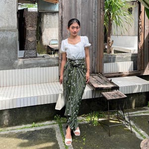 May include: A woman wearing a white blouse with a square neckline and a green and white patterned skirt. She is standing in front of a wooden door and a white tiled bench.