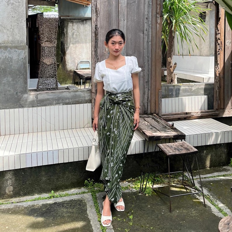 May include: A woman wearing a white blouse with a square neckline and a green and white patterned skirt. She is standing in front of a wooden door and a white tiled bench.