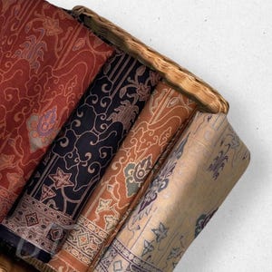 May include: Four folded batik fabrics with floral and geometric patterns are arranged in a woven basket. The fabrics are in shades of red, black, brown, and beige. The basket is made of light brown woven material.