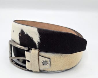 May include: A cowhide belt with a black and white pattern. The belt has a silver-colored buckle and a beige leather strap. The belt is a stylish accessory for any outfit.