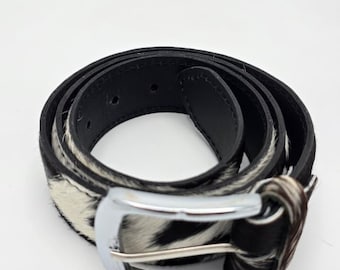 May include: A black and white cowhide belt with a silver-colored buckle. The belt is made of leather and has a classic design. The belt is approximately 1.25 inches wide.