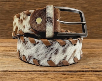 May include: A brown and white cowhide belt with a silver buckle. The belt features brown leather accents and stitching. The buckle is a dark silver color. The belt is displayed on a wooden surface.