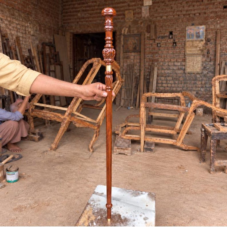 May include: A tall, slender wooden walking stick with a polished brown finish. The handle features a rounded, carved top with intricate decorative patterns and a smooth, cylindrical shaft that tapers toward a small metal tip at the base.