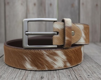 May include: A brown and white cowhide belt with a silver-colored buckle. The belt is rolled up, showcasing the texture and pattern of the hide. The buckle is a simple, rectangular design.