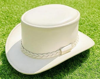 May include: A solid off-white hat with a flat top and a wide brim. The hat features a decorative braided band encircling the base of the crown, which is accented by a small, rectangular, vertically ribbed metallic detail on the front side.