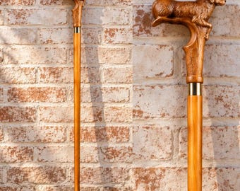 May include: A wooden walking cane featuring a carved handle in the shape of a dog lying down. The handle is connected to a smooth, light brown wooden shaft by a gold-colored metal collar. The base of the cane has a small metal tip.