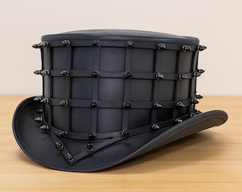 May include: A black top hat featuring a grid of thin black straps wrapped around the crown. Each intersection of the grid is adorned with a small, pointed black metal stud. The hat has a wide, curved brim and a smooth, matte black finish.