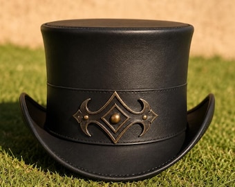 May include: A black top hat featuring a flat crown and a curved brim. A wide black band encircles the base of the crown, adorned with a central metallic bronze-toned decorative emblem shaped like a diamond with ornate, symmetrical side flourishes.