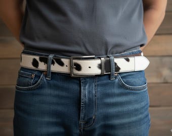 May include: A white and black cowhide belt with a silver buckle is worn over blue jeans. The belt has a rectangular silver buckle and a white leather backing. The cowhide pattern features irregular black spots.