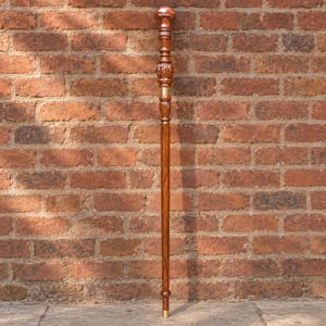 May include: A tall, slender walking stick crafted from polished brown wood with a visible grain. The handle features a rounded, carved top section with intricate detailing and a smooth, cylindrical shaft accented by a metallic gold-colored band.